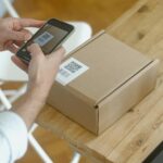 A person uses a smartphone to scan a QR code on a package in an online store setting.