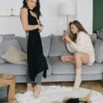 Two women relaxing while trying on new clothing purchases in a cozy living room setting.